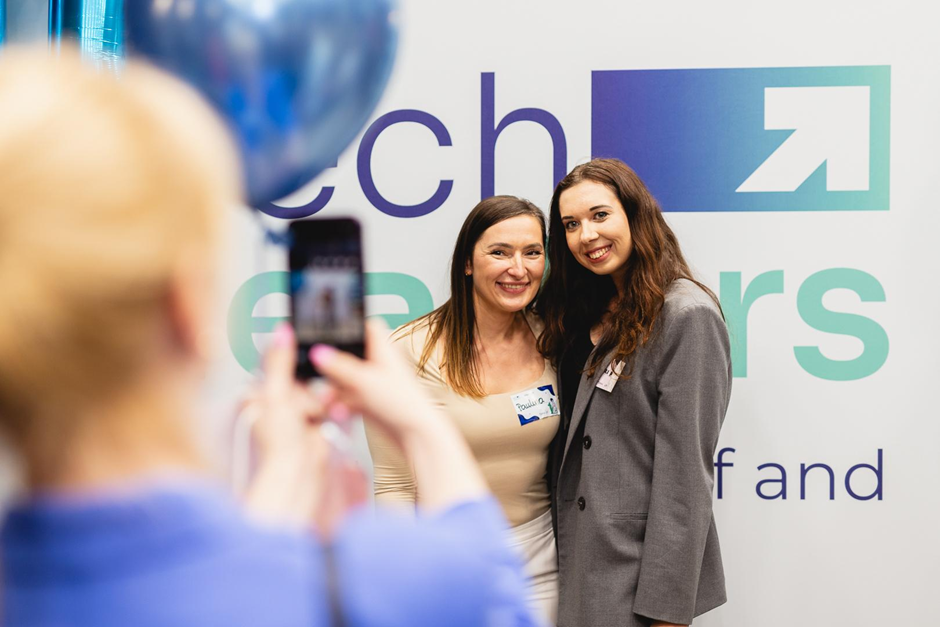 Dwie kobiety uśmiechają się do zdjęcia na tle ścianki z logo wydarzenia; na pierwszym planie osoba robi im zdjęcie smartfonem, obok widoczne niebieskie balony.