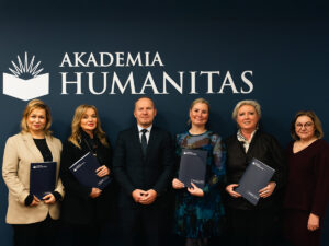 Grupowe zdjęcie sześciu osób stojących przed ścianką z logo Akademii Humanitas; uczestnicy trzymają teczki z logo uczelni, w centrum mężczyzna w garniturze.