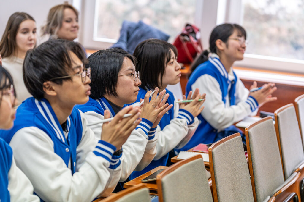 Kilka osób siedzi w ławkach sali wykładowej i klaszcze, patrząc w stronę prowadzącego; mają na sobie niebieskie bluzy z białymi rękawami.
