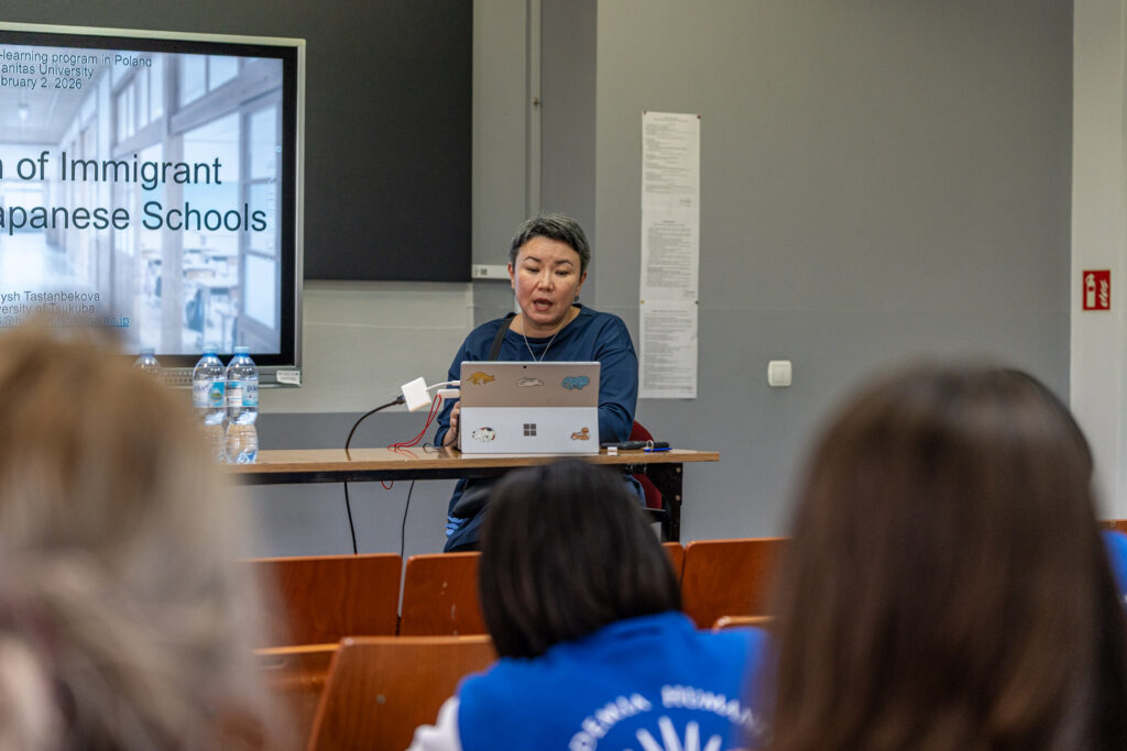 Kobieta siedzi przy stole i prowadzi prezentację, korzystając z laptopa ustawionego przed nią. Obok stoją butelki z wodą, a laptop jest podłączony do monitora wyświetlającego slajd w języku angielskim dotyczący edukacji imigrantów w Japonii.