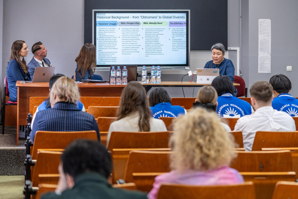 Widok sali wykładowej z perspektywy publiczności. Przy stole z przodu siedzą cztery osoby prowadzące spotkanie, w tym kobieta prezentująca z laptopa. Na ekranie wyświetlony jest slajd w języku angielskim dotyczący historycznego tła migracji i różnorodności.