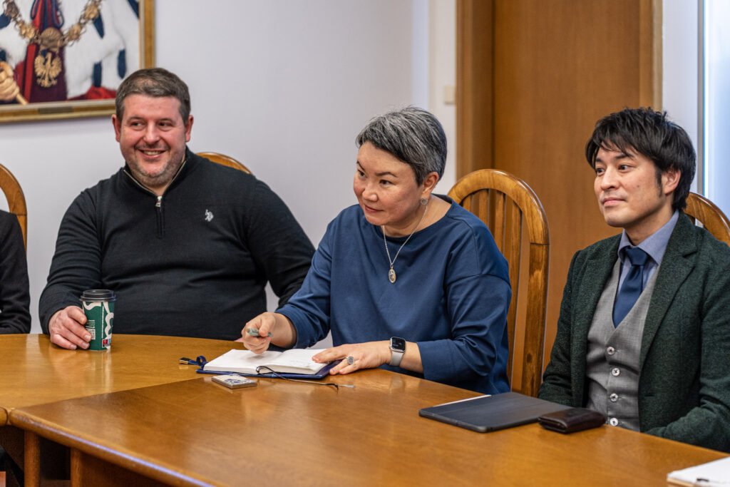 Trzy osoby siedzą przy drewnianym stole podczas spotkania. Kobieta w granatowej bluzce notuje coś w otwartym notesie, trzymając długopis w dłoni. Po jej lewej stronie siedzi uśmiechnięty mężczyzna w ciemnym swetrze, trzymający kubek z kawą na wynos.