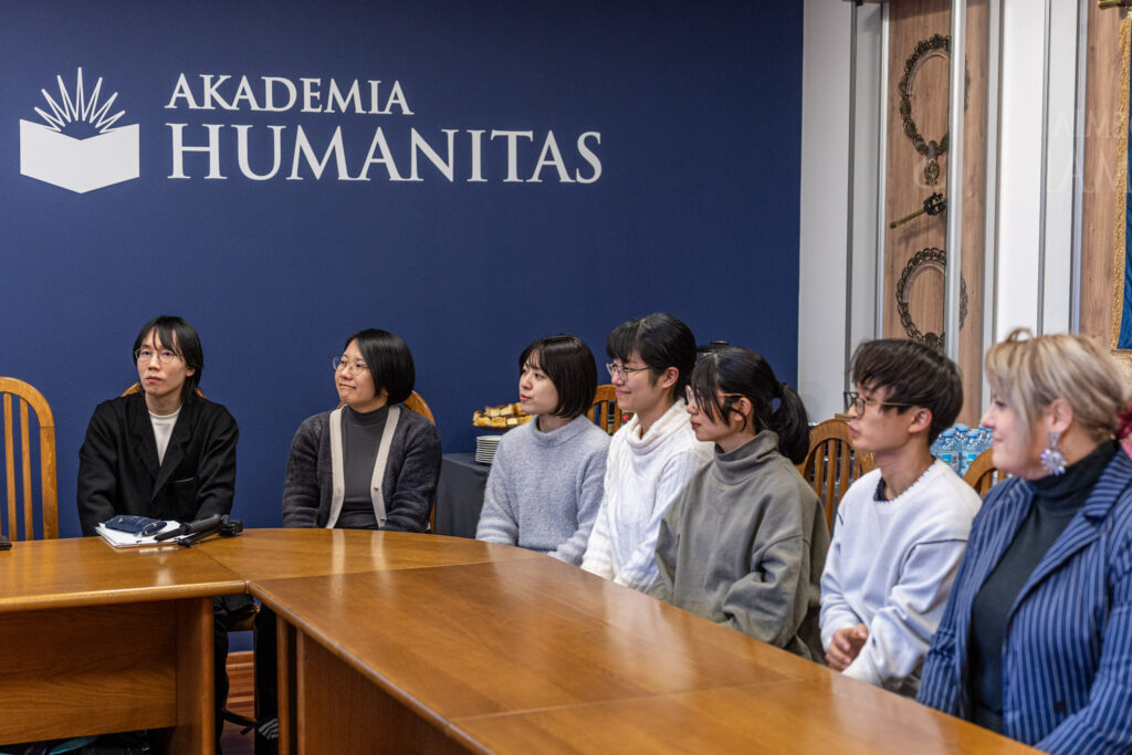 Grupa sześciu osób siedzi przy drewnianym stole w sali z granatową ścianą, na której widnieje napis „AKADEMIA HUMANITAS” oraz logo uczelni. Uczestnicy spotkania siedzą obok siebie i uważnie słuchają, zwróceni w stronę osoby znajdującej się poza kadrem.