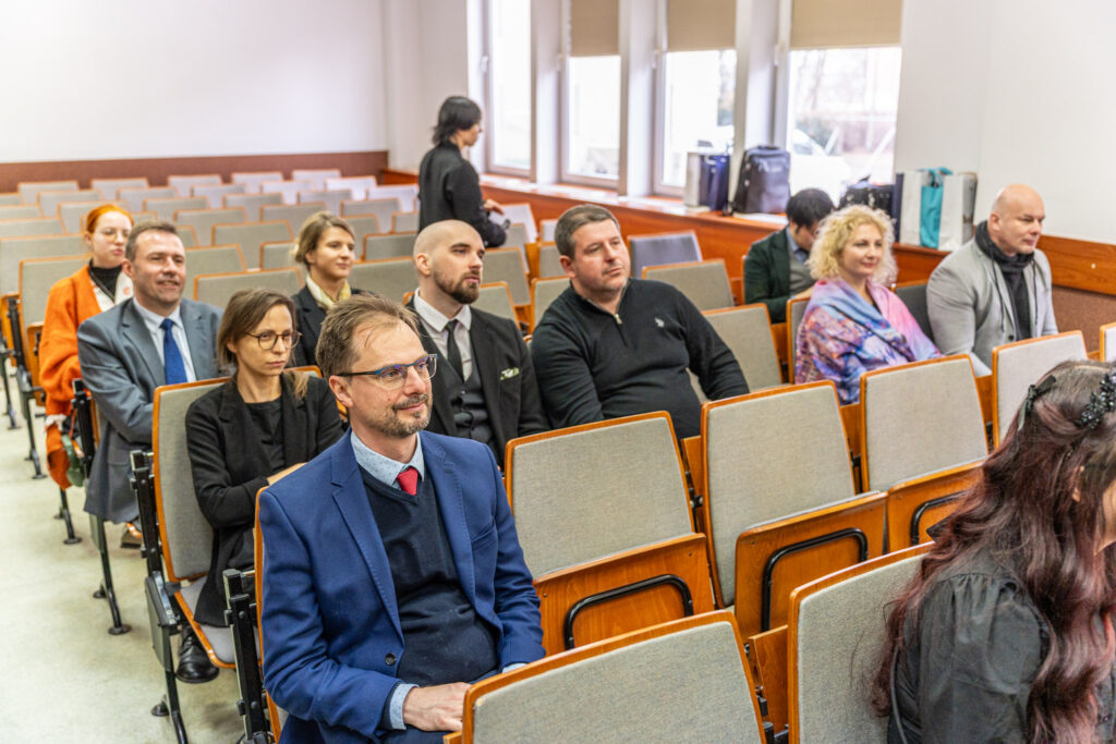Kilka osób siedzi w rzędach sali wykładowej i słucha prezentacji. Na pierwszym planie mężczyzna w niebieskiej marynarce i czerwonym krawacie siedzi skupiony, obok niego kobieta w okularach. W kolejnych rzędach widać innych uczestników, w tym osoby w garniturach i swobodniejszych strojach.