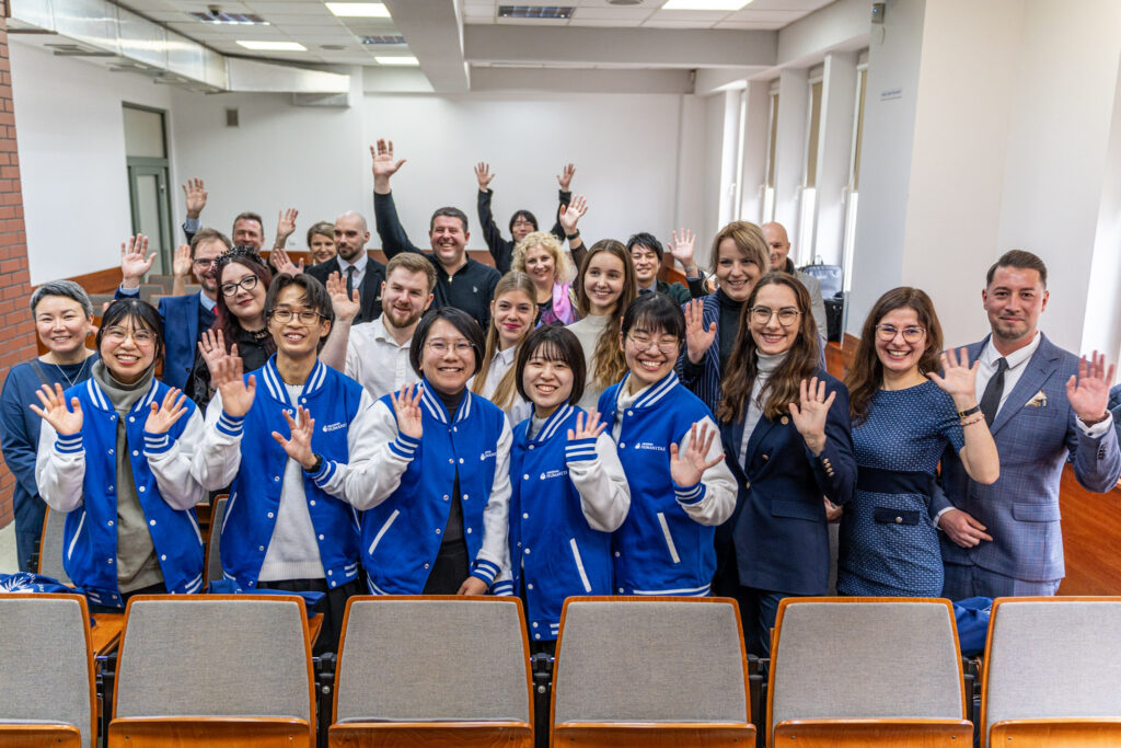 Grupa kilkunastu osób stojących w sali wykładowej uśmiecha się do zdjęcia i macha do aparatu; część uczestników ma na sobie niebieskie bluzy z logo uczelni, inni są ubrani w stroje formalne.