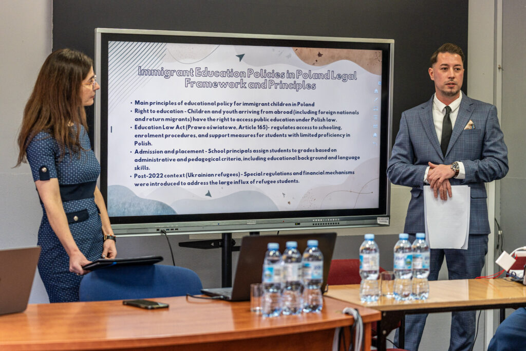 Mężczyzna w garniturze trzyma kartki i przemawia, a kobieta stoi obok z tabletem. Przed nimi znajduje się stół z laptopem i kilkoma butelkami wody.