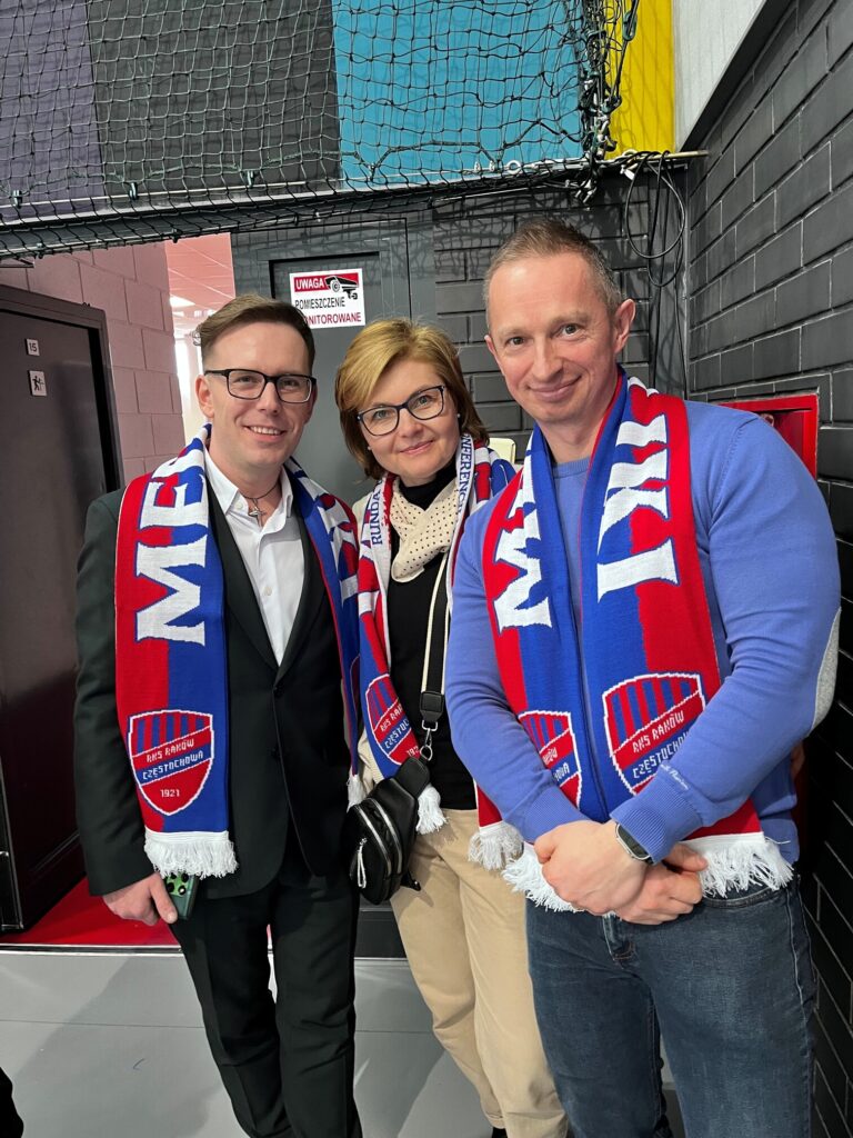Trzy osoby stoją obok siebie w hali sportowej, uśmiechnięte do obiektywu. Wszystkie mają na szyjach klubowe szaliki w barwach RKS Raków Częstochowa. W tle widoczne są ściany techniczne i siatka zabezpieczająca.
