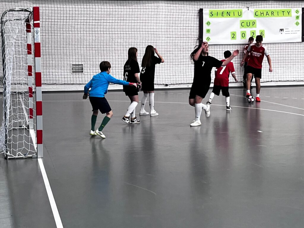 Mecz halowej piłki nożnej dzieci i młodzieży: zawodnicy w czarnych i czerwonych strojach grają pod bramką, bramkarz w niebieskiej koszulce, w tle baner „Sieniu Charity Cup 2026”