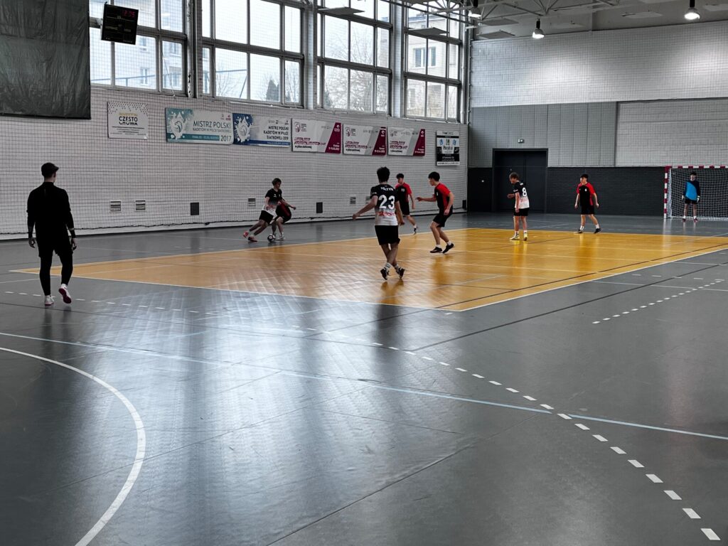 Mecz piłki halowej w hali sportowej: kilku zawodników w czarno-czerwonych strojach gra na parkiecie, w tle bramka i trybuny hali z dużymi oknami.