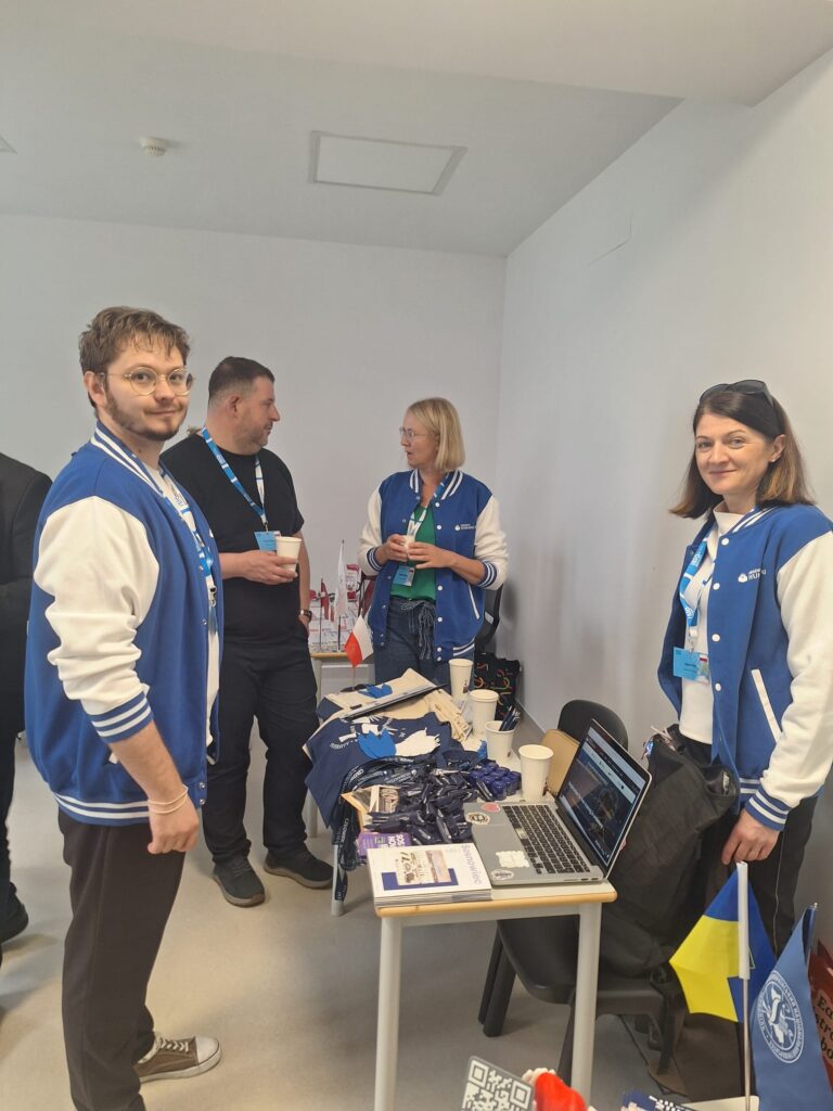 Na zdjęciu widać stoisko informacyjne podczas wydarzenia. Przy niewielkim stole stoją cztery osoby – dwie kobiety i dwóch mężczyzn – ubrane w niebiesko-białe bluzy z logo uczelni Akademia Humanitas.
Na stole znajdują się materiały promocyjne, smycze, ulotki, kubki oraz otwarty laptop. Widać także małe flagi, w tym flagę Ukrainy. Atmosfera jest swobodna, uczestnicy rozmawiają i prezentują ofertę.