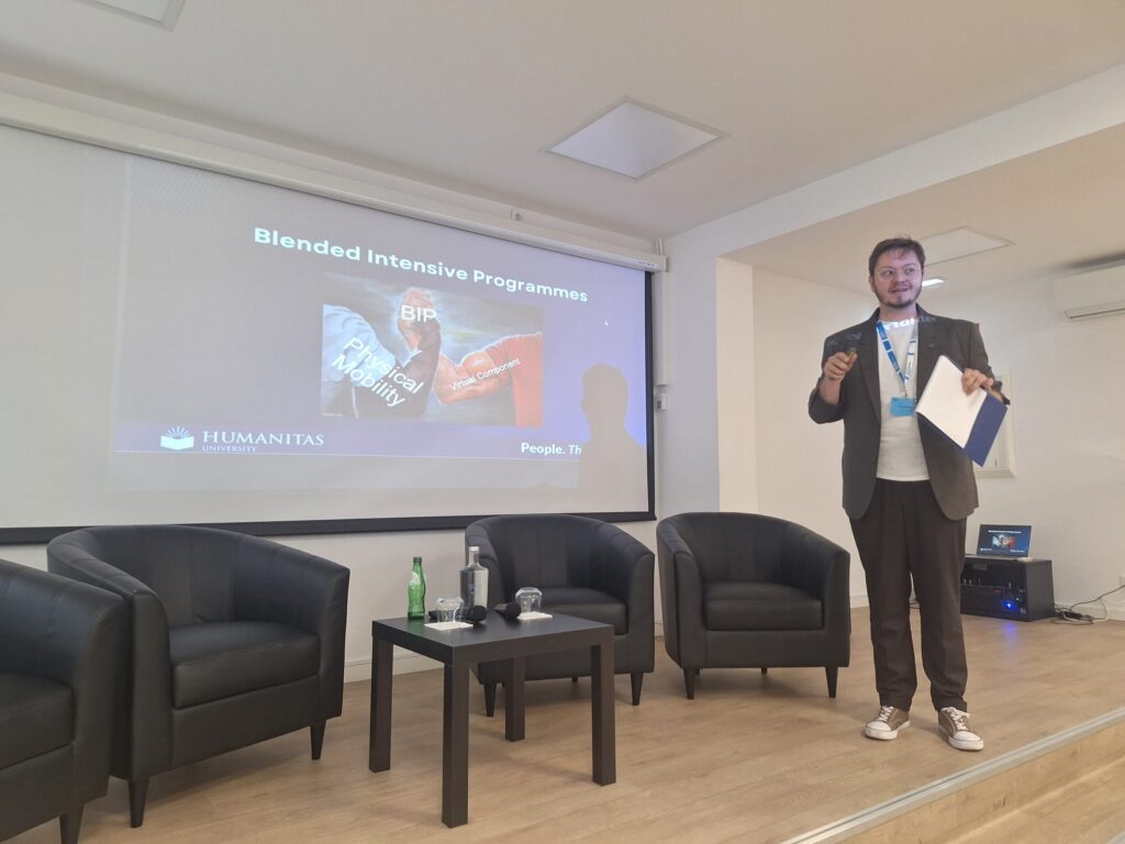 Na zdjęciu widać mężczyznę prowadzącego prezentację w sali konferencyjnej. Stoi obok ekranu, na którym wyświetlony jest slajd dotyczący programu „Blended Intensive Programmes” z logo uczelni Humanitas.
Prelegent trzyma notatki i gestykuluje, zwracając się do uczestników. Obok ustawione są fotele i stolik z wodą, co sugeruje przygotowanie do panelu lub dyskusji.