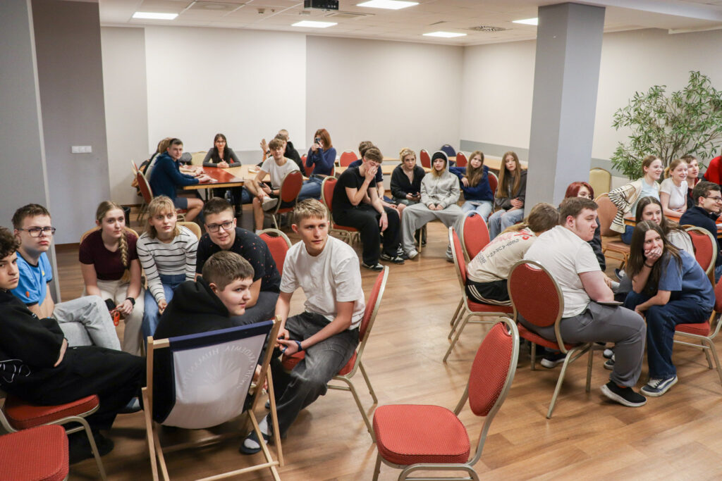 Grupa młodzieży siedzi przy stolikach w sali warsztatowej, rozmawia i uczestniczy w zajęciach integracyjnych.