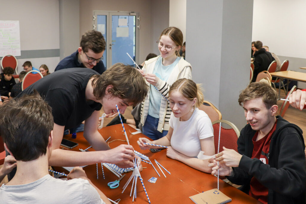 Grupa młodzieży siedzi przy stole i wspólnie buduje konstrukcję ze słomek podczas warsztatów integracyjnych.