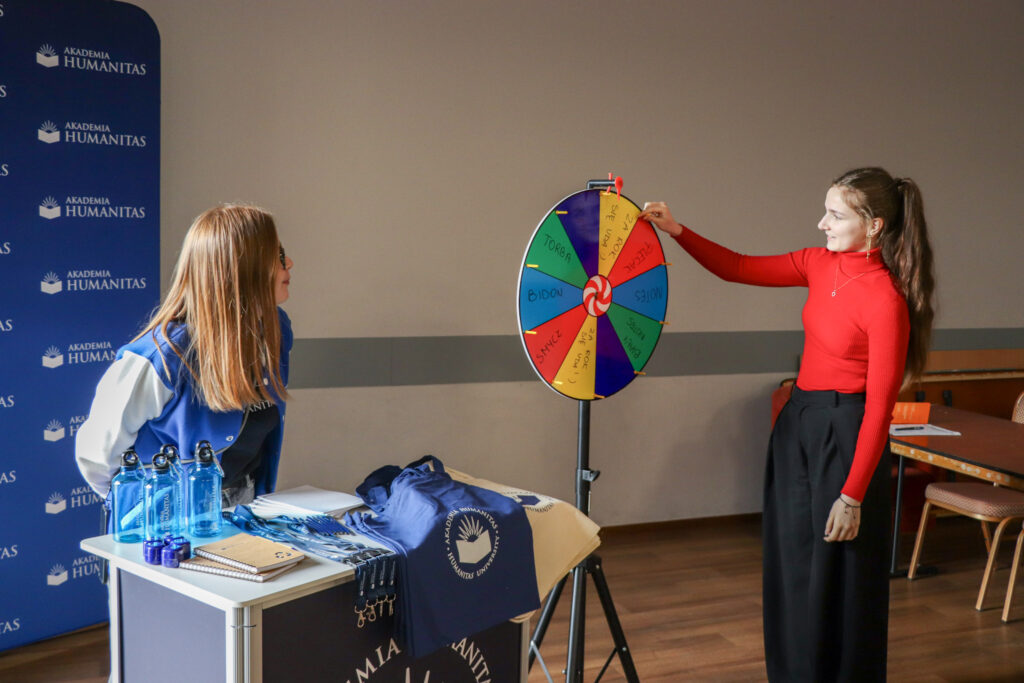 Uczennica przy stoisku Akademii Humanitas kręci kołem z nagrodami podczas wydarzenia.