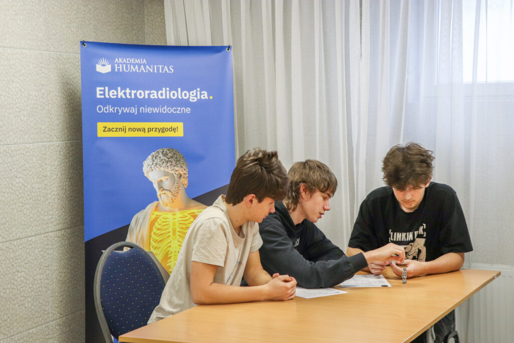 Uczniowie pracują zespołowo przy zadaniu edukacyjnym podczas warsztatów. W tle baner kierunki Elektroradiologia Akademii Humanitas. 