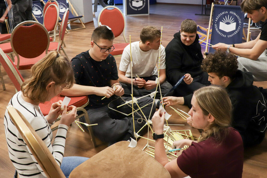 Grupa młodzieży siedzi na podłodze i wspólnie buduje konstrukcję ze słomek podczas warsztatów integracyjnych.