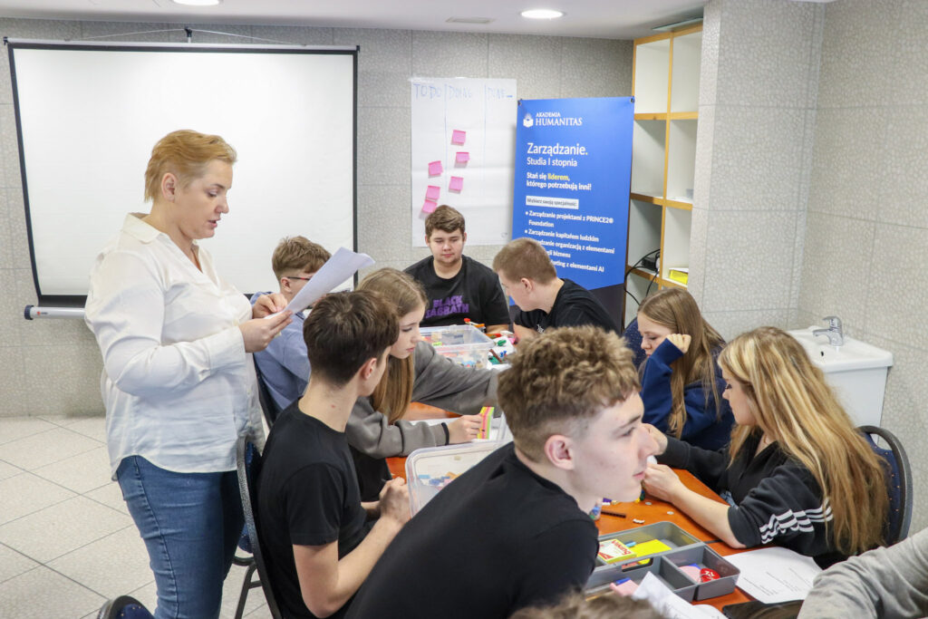 Prowadząca czyta instrukcję, a grupa młodzieży pracuje przy stole nad zadaniem zespołowym podczas warsztatów w Akademii Humanitas.