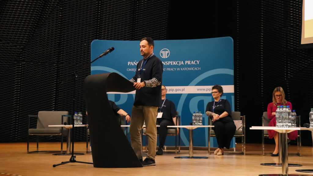 Na zdjęciu widoczna jest scena konferencyjna podczas II Śląskiej Debaty o Bezpieczeństwie Pracy w Katowicach w 2026 roku, nad którą patronat honorowy objęła Akademia Humanitas.

Na pierwszym planie stoi mężczyzna przemawiający przy mównicy z mikrofonem. Ma na sobie identyfikator konferencyjny i mówi do uczestników wydarzenia. Za nim, na scenie, siedzi kilka osób biorących udział w debacie – kobiety i mężczyźni ubrani w stroje formalne lub półformalne. Przed nimi ustawione są stoliki z butelkami wody.