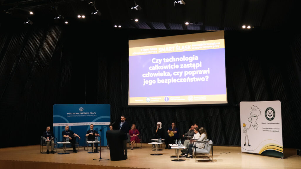 Na zdjęciu widać scenę podczas debaty o bezpieczeństwie pracy. Na środku stoi mężczyzna przemawiający przy mównicy, a po obu stronach siedzą uczestnicy panelu.

Nad sceną znajduje się duży ekran z pytaniem o to, czy technologia zastąpi człowieka, czy poprawi jego bezpieczeństwo. Po bokach stoją plansze z logotypami instytucji związanych z bezpieczeństwem pracy.