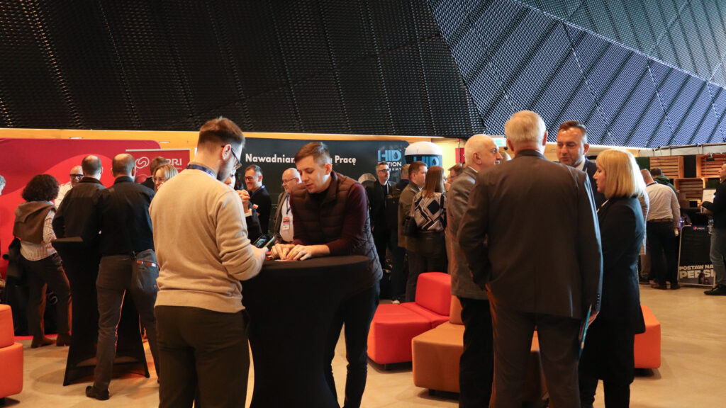 Na zdjęciu widać przestrzeń networkingową podczas wydarzenia branżowego. W dużej, nowoczesnej hali stoi wiele osób rozmawiających w małych grupach przy wysokich stolikach. Uczestnicy są ubrani w stroje biznesowe i półformalne, a część z nich ma identyfikatory konferencyjne na smyczach.

W tle znajdują się stoiska firm i sponsorów z widocznymi logotypami oraz elementy strefy wypoczynku z czerwonymi i pomarańczowymi siedziskami. Atmosfera przypomina przerwę między panelami dyskusyjnymi, kiedy uczestnicy wymieniają się kontaktami i rozmawiają o wydarzeniu.