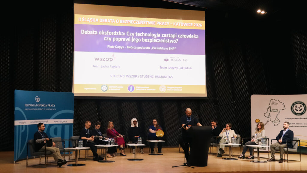 Na zdjęciu widać scenę podczas II Śląskiej Debaty o Bezpieczeństwie Pracy w Katowicach w 2026 roku, nad którą patronat honorowy objęła Akademia Humanitas.

Na środku stoi mężczyzna przemawiający przy mównicy, a po obu stronach sceny siedzą uczestnicy debaty. Za nimi widoczny jest duży ekran z tytułem wydarzenia oraz temat debaty oksfordzkiej o wpływie technologii na bezpieczeństwo pracy. Po bokach znajdują się plansze z logotypami instytucji, w tym Państwowej Inspekcji Pracy.
