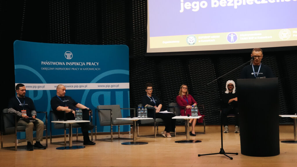 Na zdjęciu widać scenę podczas II Śląskiej Debaty o Bezpieczeństwie Pracy w Katowicach w 2026 roku, nad którą patronat honorowy objęła Akademia Humanitas.

Na środku stoi mężczyzna przemawiający przy mównicy, a po obu stronach sceny siedzą uczestnicy debaty. Za nimi widoczny jest duży ekran z tytułem wydarzenia oraz temat debaty oksfordzkiej o wpływie technologii na bezpieczeństwo pracy. Po bokach znajdują się plansze z logotypami instytucji, w tym Państwowej Inspekcji Pracy.