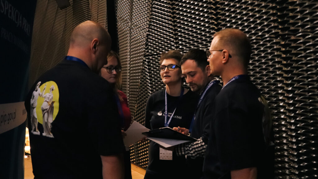 Na zdjęciu widać drużynę Akademii Humanitas stojącą razem za kulisami sceny podczas II Śląskiej Debaty o Bezpieczeństwie Pracy w Katowicach w 2026 roku, nad którą patronat honorowy objęła Akademia Humanitas.

Uczestnicy mają na sobie identyfikatory konferencyjne i koszulki z logo uczelni. Rozmawiają i omawiają materiały zapisane na kartkach oraz na tablecie, prawdopodobnie przygotowując się do kolejnej części debaty. Jedna z osób trzyma dokumenty, a inna spogląda na ekran urządzenia.