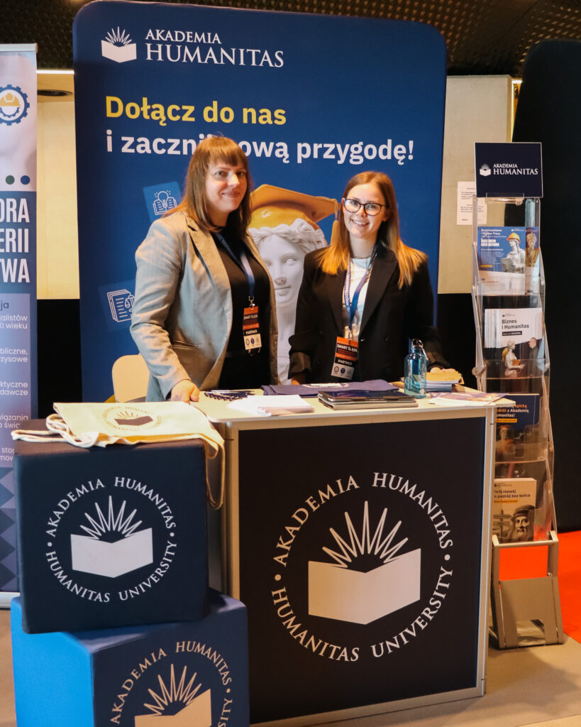 Na zdjęciu widać stoisko promocyjne Akademii Humanitas podczas wydarzenia. Za ladą stoją dwie kobiety z identyfikatorami konferencyjnymi i uśmiechają się do zdjęcia.

Za nimi znajduje się duży baner z napisem „Dołącz do nas i zacznij nową przygodę!”. Na stoliku leżą materiały promocyjne uczelni, a obok stoi stojak z broszurami i ulotkami.