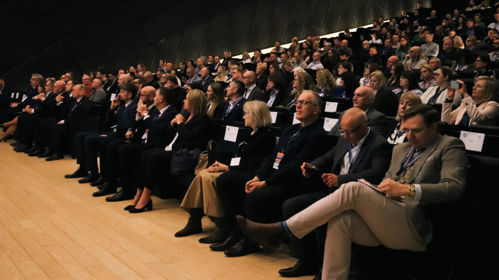 Na zdjęciu widać dużą salę audytoryjną wypełnioną uczestnikami konferencji. Kilkadziesiąt osób siedzi w rzędach na podwyższonych miejscach i słucha wystąpienia odbywającego się poza kadrem.

Większość uczestników ma na sobie stroje biznesowe oraz identyfikatory konferencyjne. Niektórzy patrzą w stronę sceny, inni robią notatki lub sprawdzają telefon. Atmosfera jest skupiona i oficjalna, typowa dla panelu lub wykładu podczas wydarzenia branżowego.