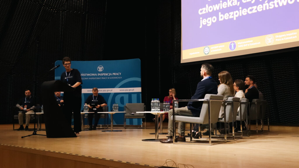 Na zdjęciu widać scenę podczas II Śląskiej Debaty o Bezpieczeństwie Pracy w Katowicach w 2026 roku, nad którą patronat honorowy objęła Akademia Humanitas.

Na środku stoi kobieta przemawiająca przy mównicy, a po obu stronach sceny siedzą uczestnicy debaty. Za nimi widoczny jest duży ekran z tytułem wydarzenia oraz temat debaty oksfordzkiej o wpływie technologii na bezpieczeństwo pracy. Po bokach znajdują się plansze z logotypami instytucji, w tym Państwowej Inspekcji Pracy.