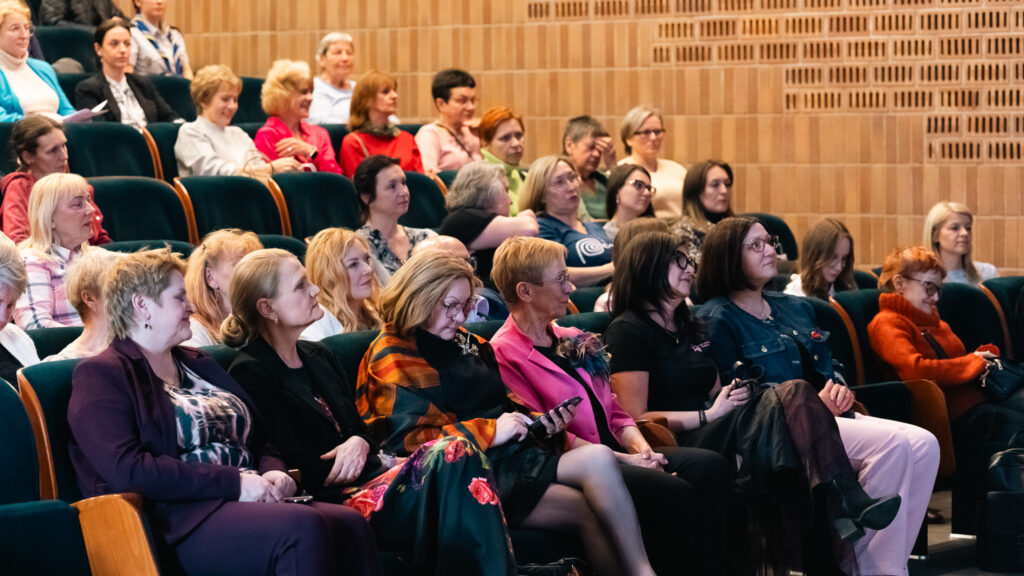 Publiczność siedzi w auli i uważnie słucha wydarzenia. Uczestnicy są skupieni, część notuje lub korzysta z telefonów.