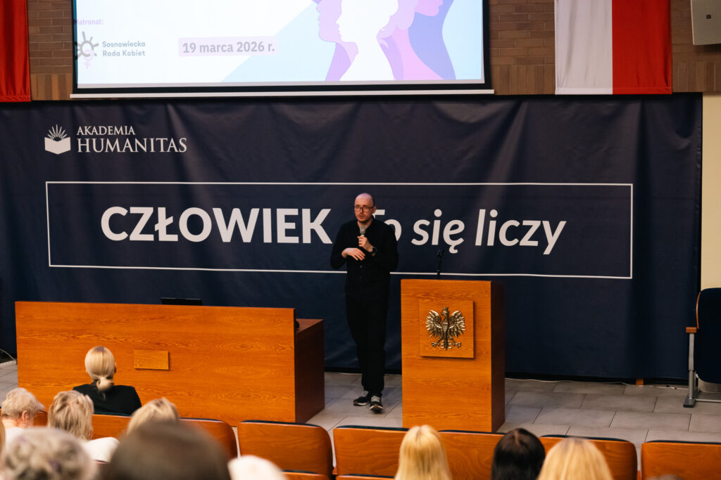 Prelegent przemawia do publiczności w auli Akademii Humanitas na tle hasła „Człowiek się liczy”. Uczestnicy siedzą na widowni i słuchają wystąpienia.