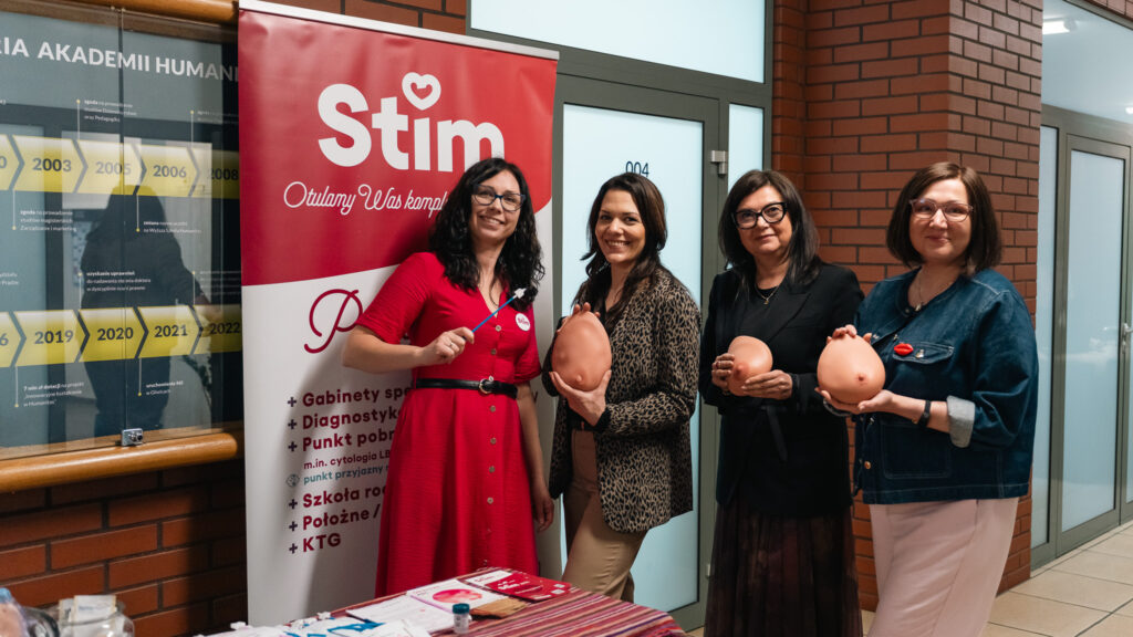 Cztery kobiety stoją przy stoisku promocyjnym i prezentują materiały edukacyjne. Trzymają modele i uśmiechają się do zdjęcia.