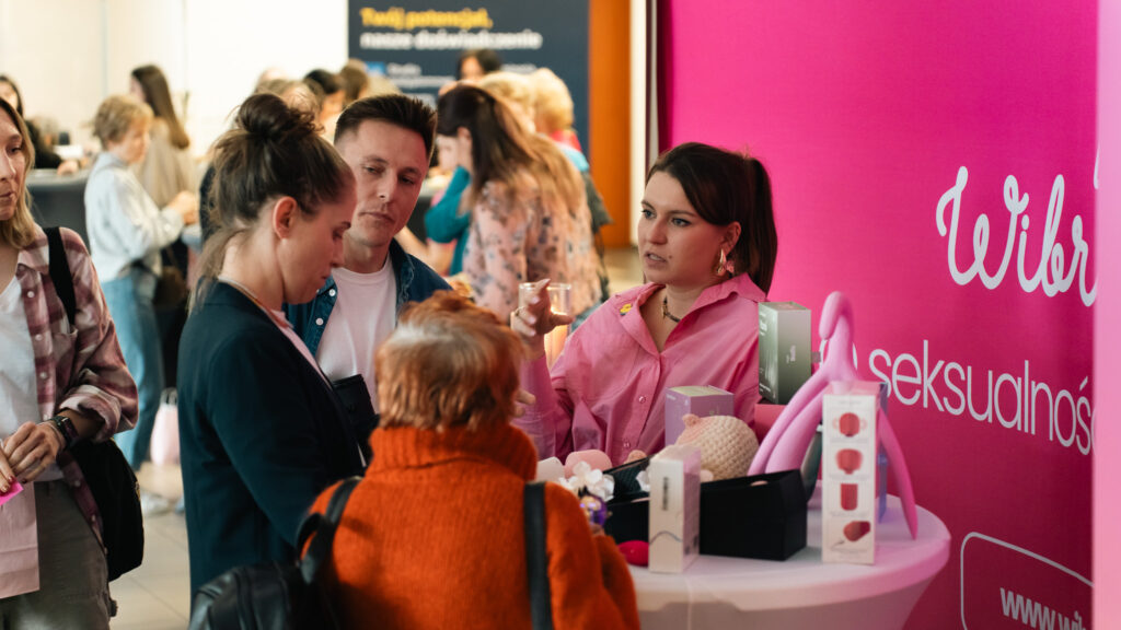 Uczestnicy odwiedzają stoisko edukacyjne dotyczące seksualności, gdzie prowadząca prezentuje produkty i materiały. Wokół trwa rozmowa i zainteresowanie ofertą.