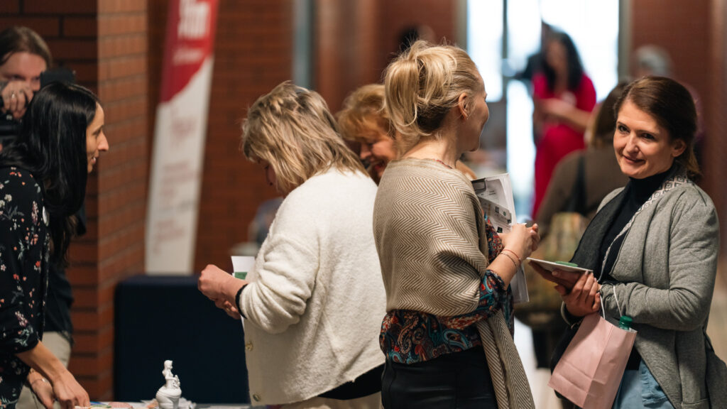 Grupa kobiet rozmawia w przestrzeni wydarzenia, trzymając materiały i drobne upominki. Atmosfera jest swobodna i sprzyja wymianie doświadczeń.