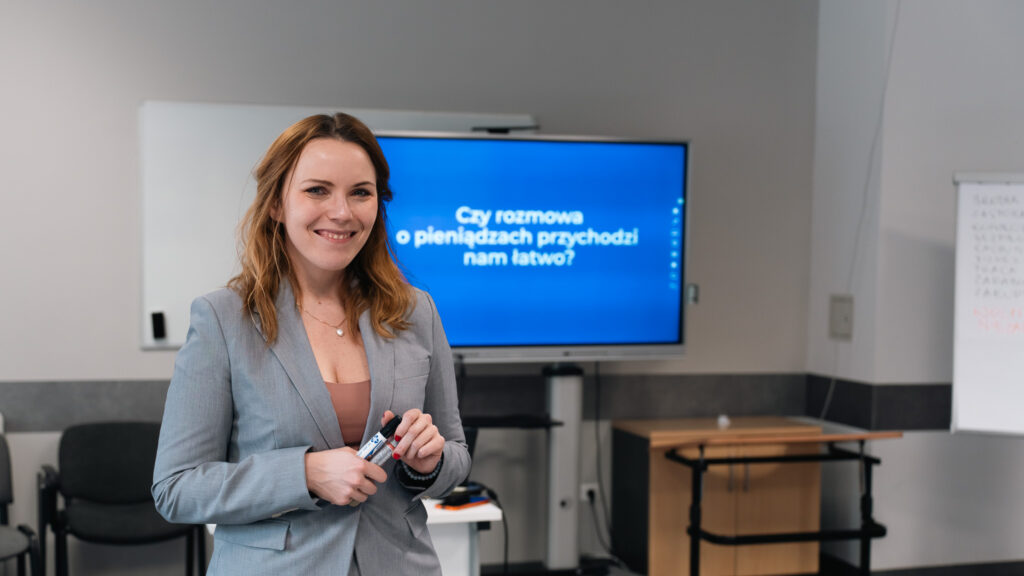 Kobieta prowadzi szkolenie w sali dydaktycznej, stojąc przed ekranem z prezentacją. Uczestnicy siedzą naprzeciwko i słuchają.