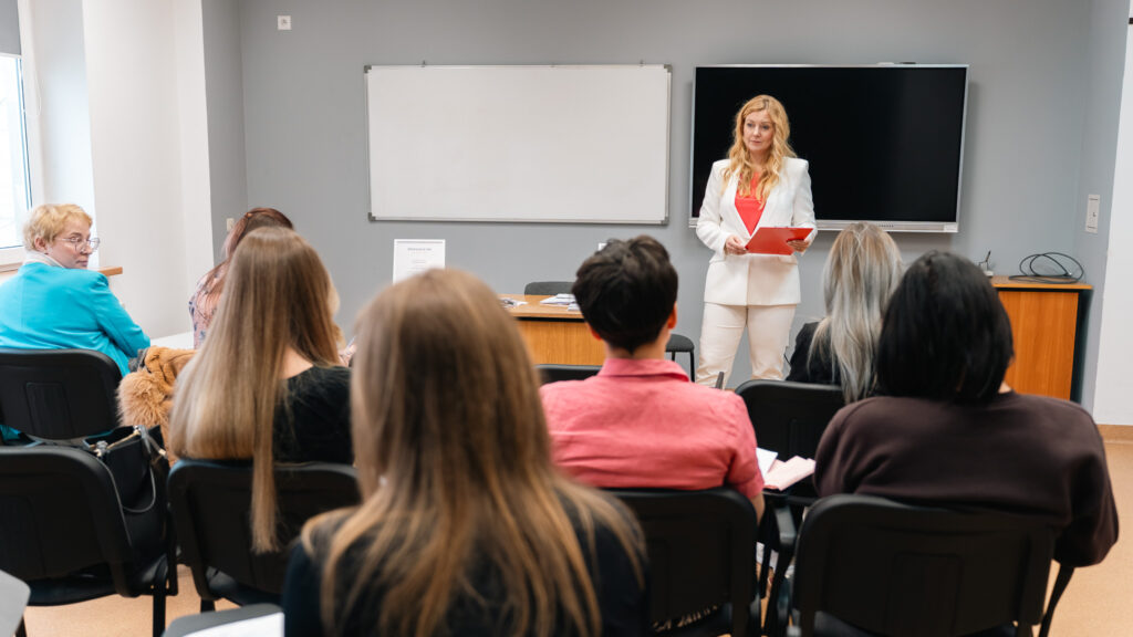 Prowadząca w jasnym stroju prezentuje materiały przed grupą słuchaczy w sali szkoleniowej. Uczestnicy siedzą w rzędach i skupiają się na wystąpieniu.