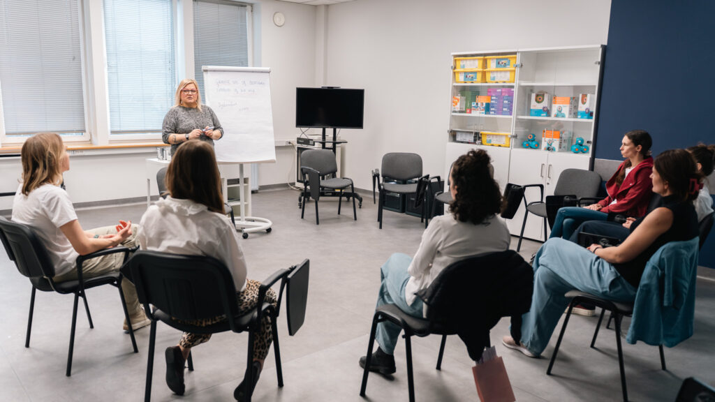 Trenerka prowadzi zajęcia warsztatowe w kręgu, stojąc przy flipcharcie. Uczestnicy siedzą wokół i biorą udział w dyskusji.
