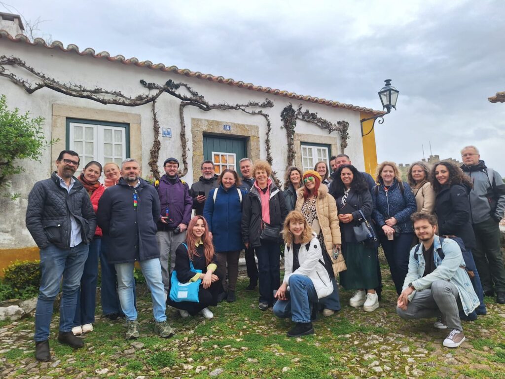 Na zdjęciu widać grupę kilkunastu osób stojących razem na zewnątrz, przed niewielkim, białym domem z zielonymi okiennicami i drzwiami.
Uczestnicy są ubrani w kurtki, co sugeruje chłodniejszą pogodę. Kilka osób kuca z przodu, reszta stoi w rzędzie, uśmiechając się do zdjęcia. W tle widać rośliny pnące się po ścianie budynku oraz pochmurne niebo.