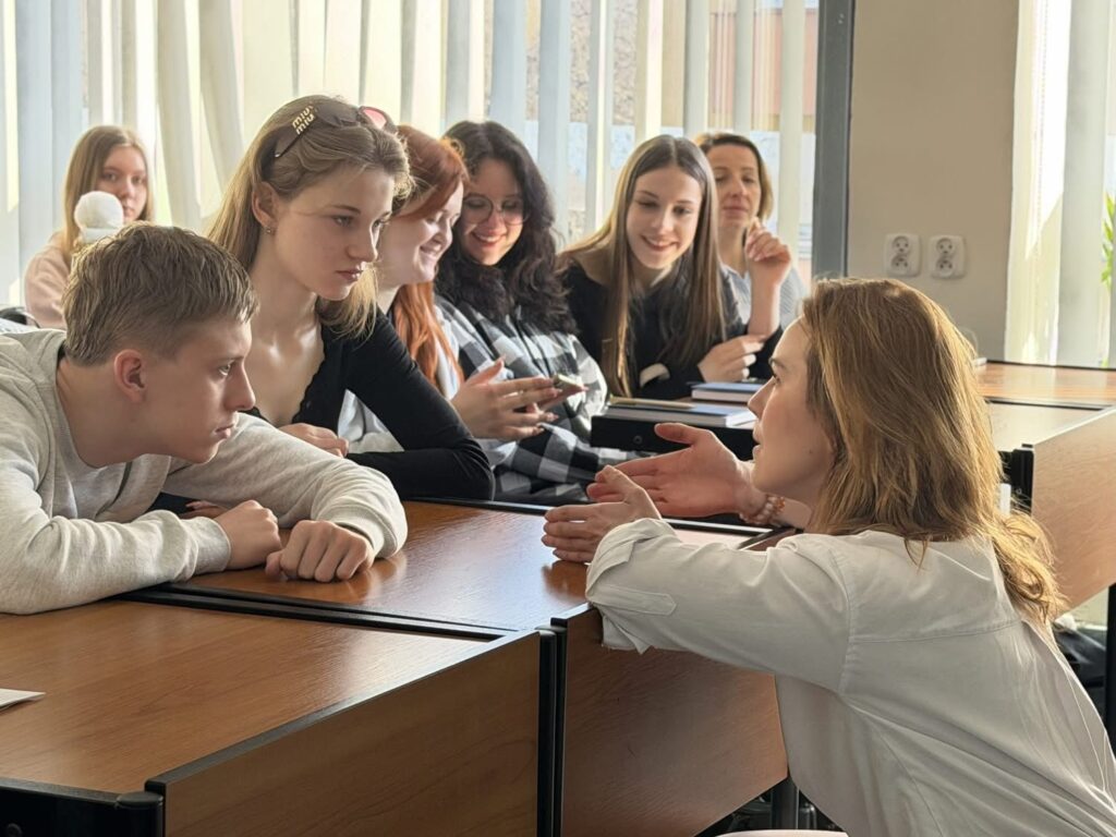 Na zdjęciu widać grupę młodych osób siedzących w sali wykładowej. Kilkoro studentów siedzi przy ławkach, a prowadząca zajęcia nachyla się i rozmawia z nimi, gestykulując rękami.