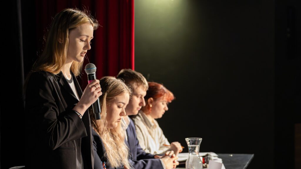 Uczestniczka przemawia do mikrofonu, a pozostali przy stole słuchają i notują.