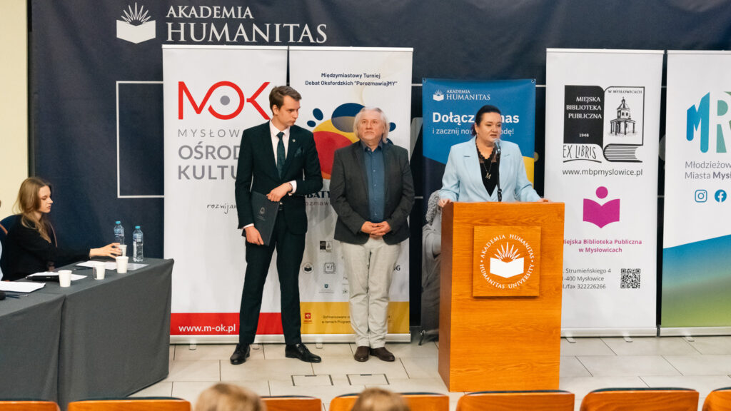 Kobieta przemawia przy mównicy Akademii Humanitas, obok stoją dwaj mężczyźni, a w tle widać banery organizatorów i partnerów wydarzenia.