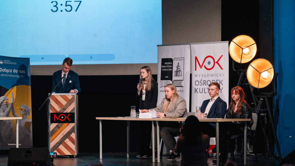 Debata na scenie – jedna osoba przemawia, inni uczestnicy siedzą przy stole, a moderator stoi przy mównicy.