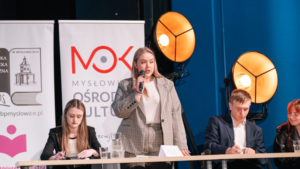 Dziewczyna przemawia do mikrofonu podczas debaty; obok siedzą uczestnicy i robią notatki.
