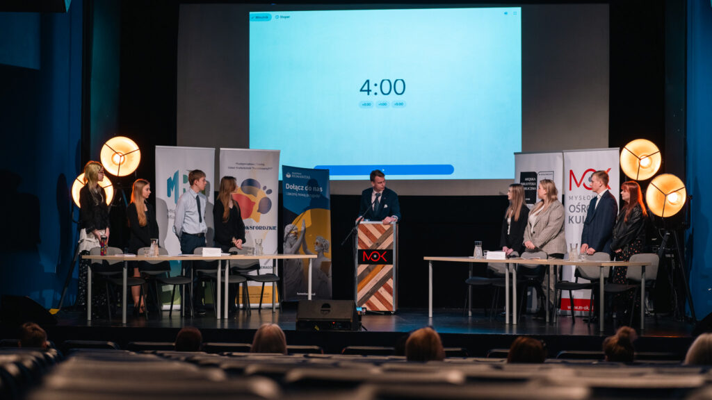 Debata na scenie: dwie drużyny stoją przy stołach, moderator przy mównicy, a na ekranie w tle widać licznik czasu.