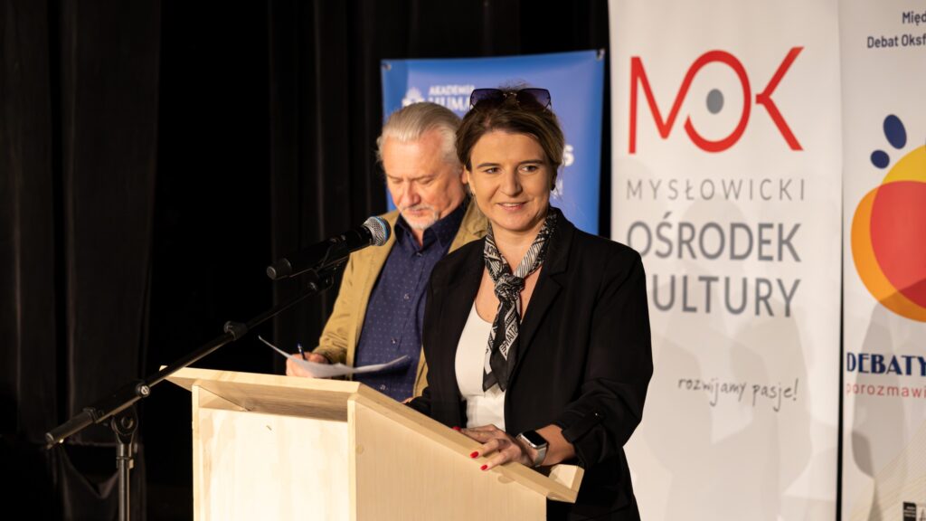 Kobieta przemawia przy mównicy, a za nią stoi mężczyzna z notatkami.