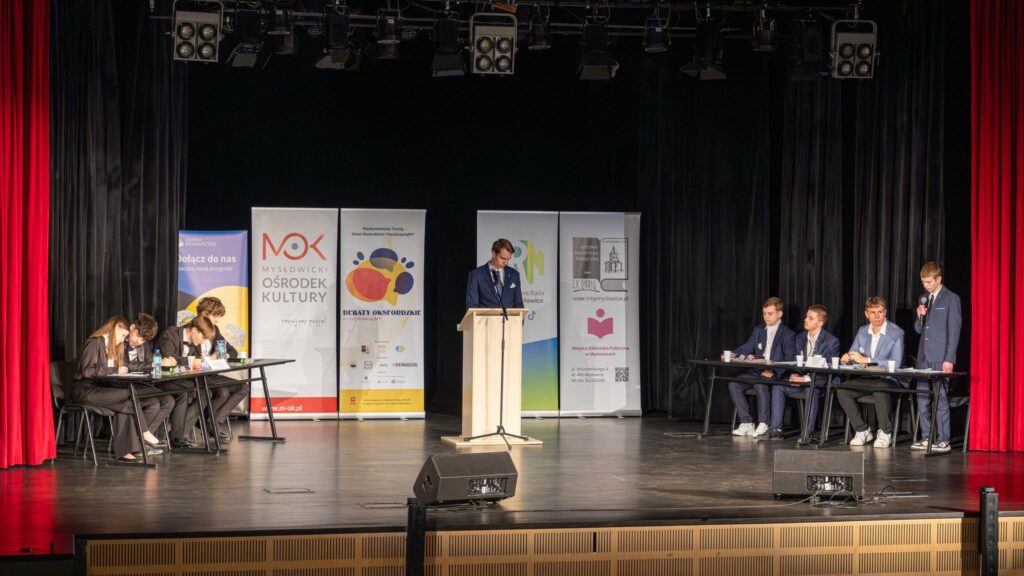 Debata na scenie – dwie drużyny przy stołach, mówca przy mównicy i uczestnik zabierający głos.