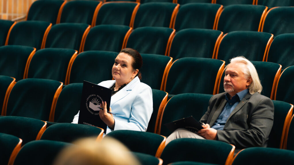 Dwie osoby siedzą na widowni w sali audytoryjnej i trzymają notatki, obserwując wydarzenie.