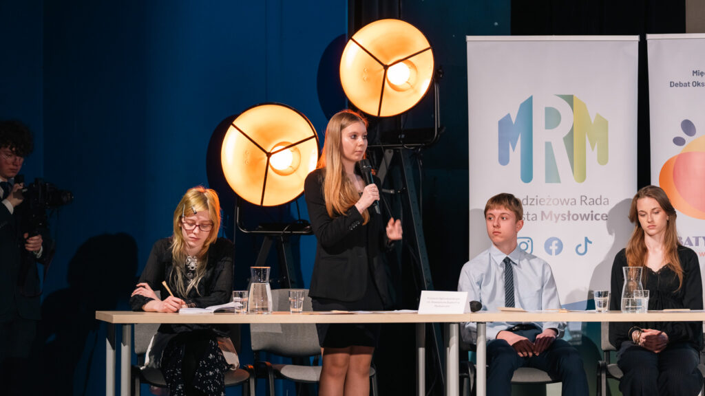 Uczennica przemawia przy stole podczas debaty; pozostali uczestnicy słuchają i notują.
