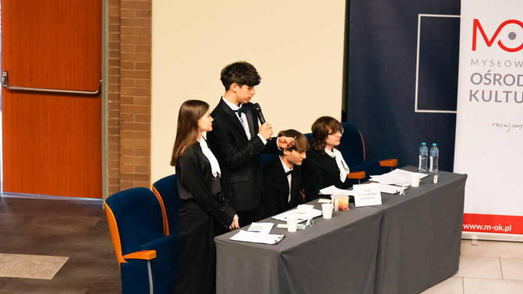 Uczeń przemawia przy stole podczas debaty; pozostali uczestnicy słuchają i notują.